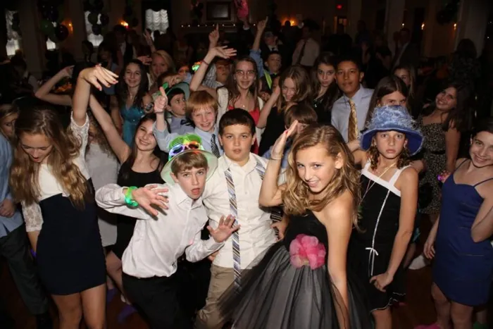 Teens in party hats and glow necklaces dancing and laughing at a mitzvah party.