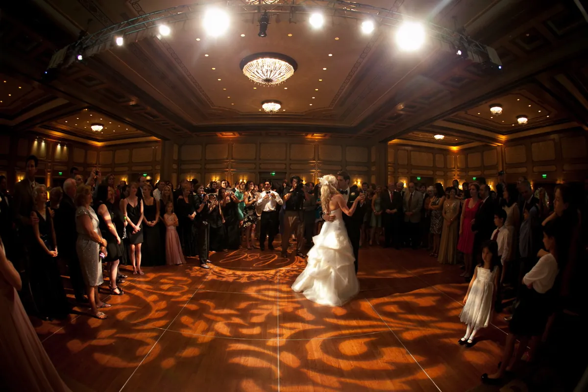 A bride dancing in a ballroom washed in warm patterned gobo light, guests standing around the floor.