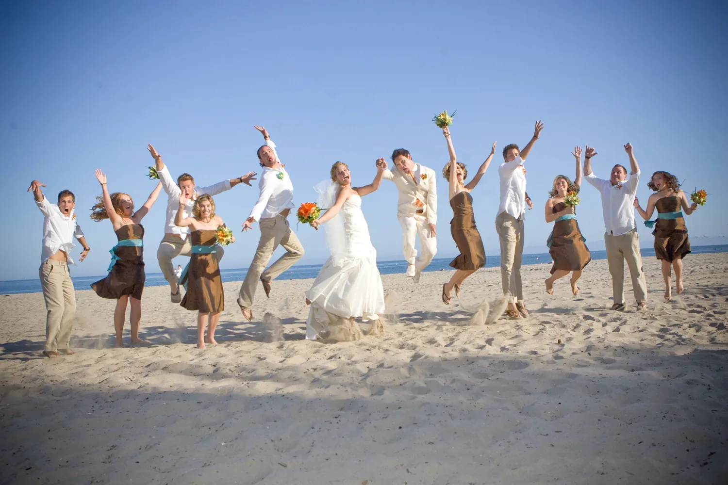 A wedding party in white and brown jumping in the air together on a sunny beach.