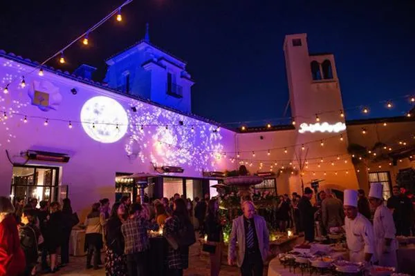 An outdoor reception at night, the building lit purple and a crowd gathered below.