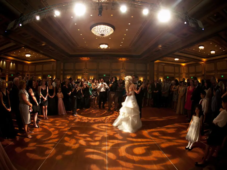 A bride and groom share their first dance in a ballroom lit with amber gobo patterns.