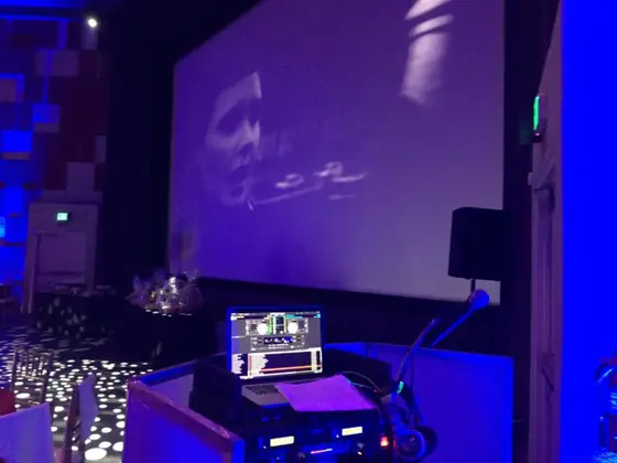 A DJ booth in front of a large projection screen in blue light.