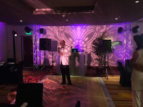 An MC at the microphone in a room framed by truss lighting and a patterned gobo wall.