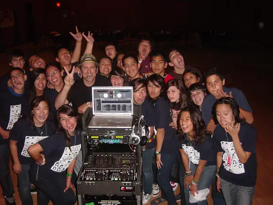 A school dance crowd posing around the DJ setup with their hands up.