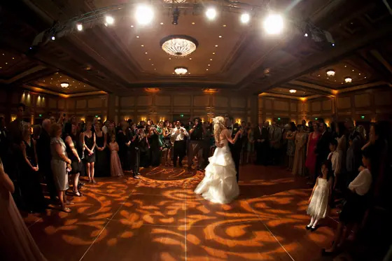 A bride and groom dance in a ballroom while patterned amber light covers the floor around them.