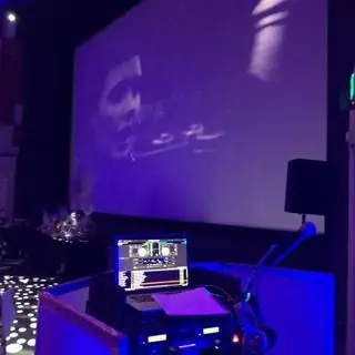 A DJ booth in front of a large projection screen in a room washed in blue light.